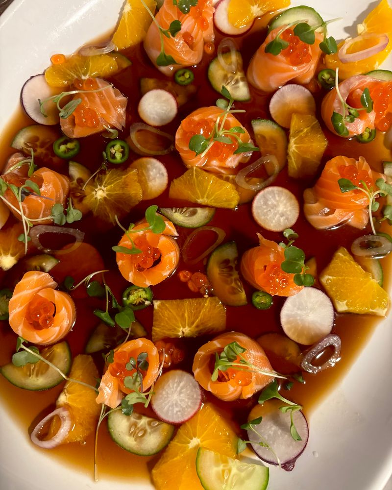 A white platter of salmon rosettes topped with salmon roe and microgreens, scattered with orange, radish, cucumber and jalapeño.